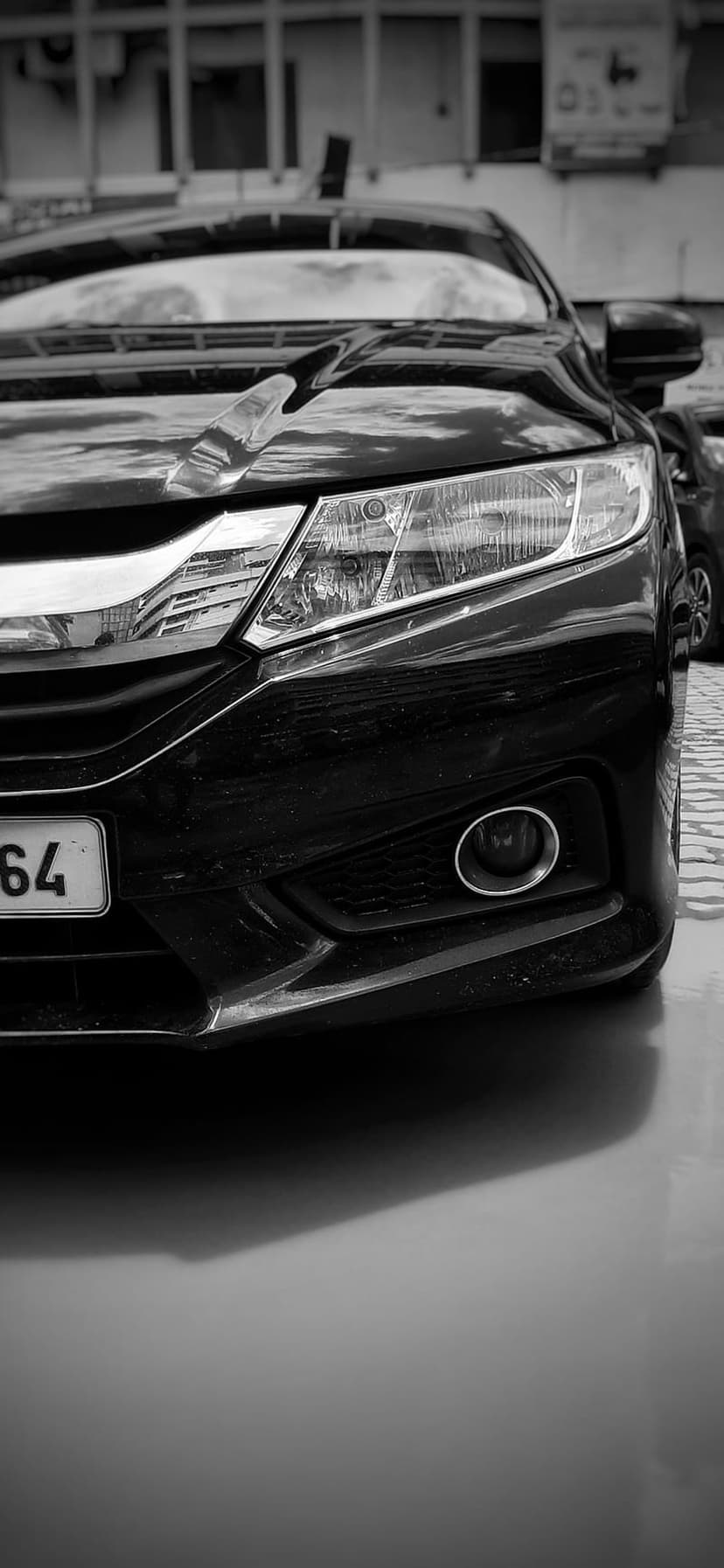 Black car headlight and bumper with building reflections in a high-contrast monochrome close-up photography style.