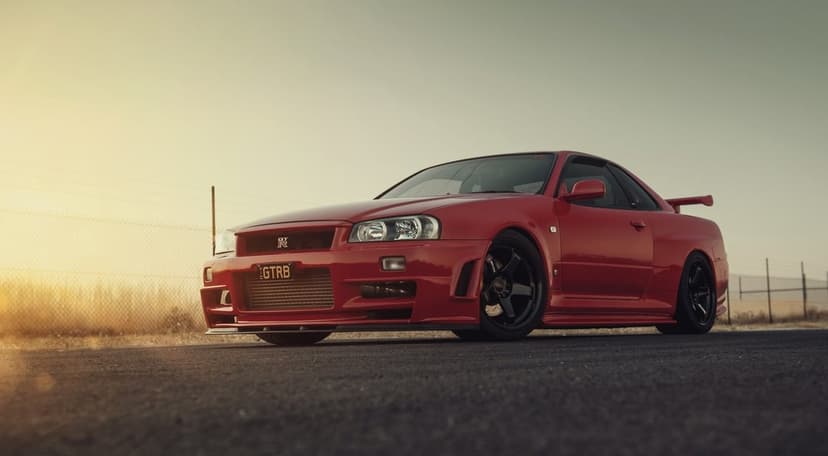 Red Nissan Skyline R34 GT-R parked on asphalt during golden hour with a soft sunset background.