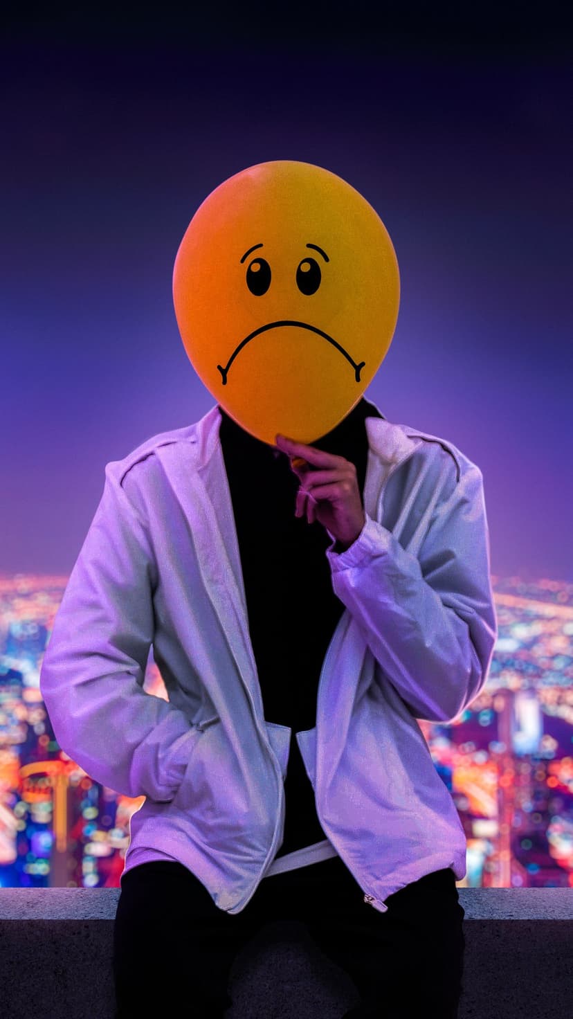 Person holding a yellow sad face balloon over their head against a blurred purple and blue city night sky.
