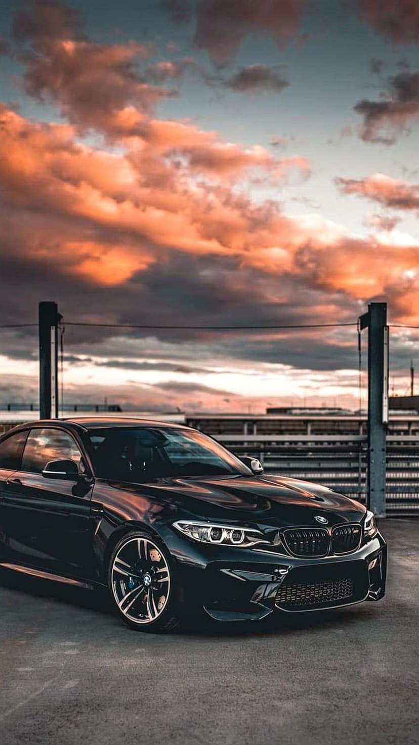 Black BMW M2 sports car with glowing headlights parked on concrete under a dramatic orange and grey sunset sky.