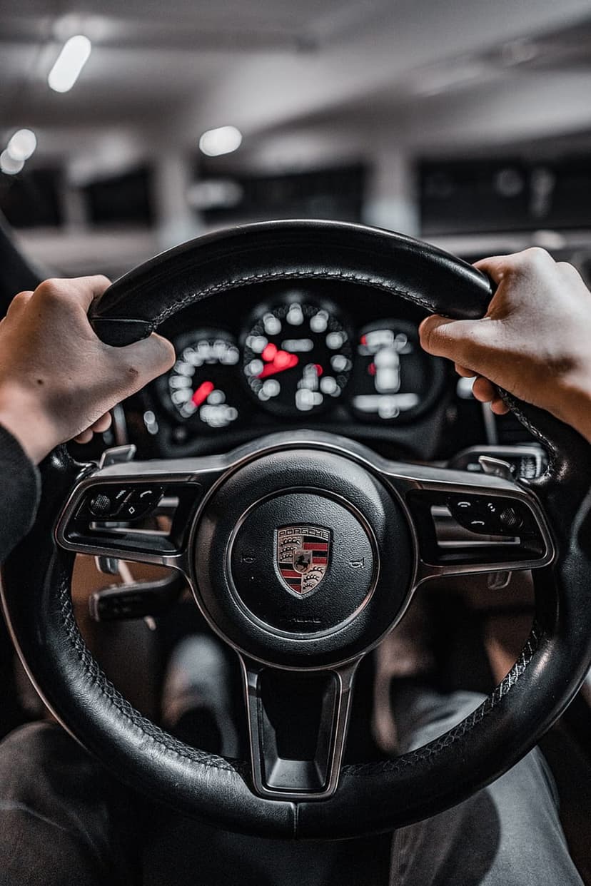 Hands grip a black leather Porsche steering wheel with a blurred luxury dashboard and glowing gauges in the back.