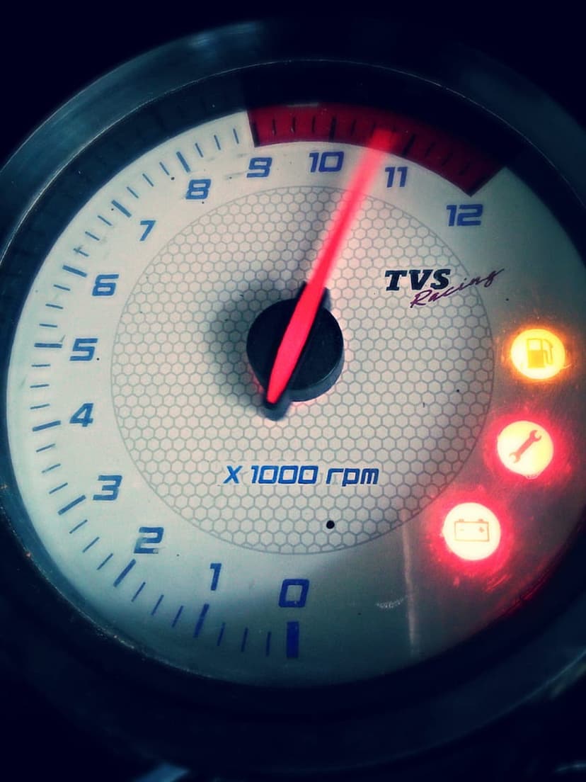 TVS Racing tachometer needle at 10,500 RPM with glowing fuel and battery icons visible on a dark dashboard.