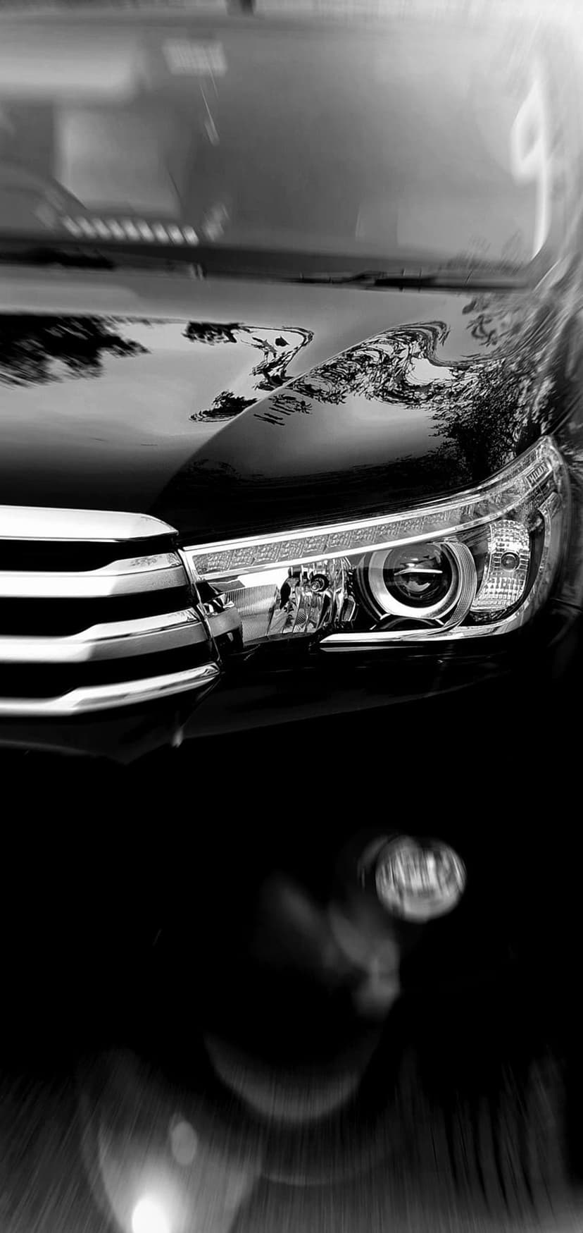 Modern car headlight and chrome grille with tree reflections and soft bokeh lights in a monochrome palette.