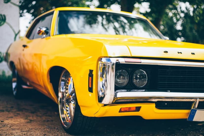 Yellow vintage muscle car with a chrome grille and dual headlights parked on gravel against blurred green trees.