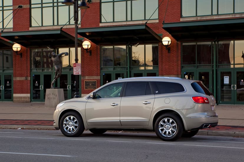 Champagne Buick Enclave SUV parked on an urban street in front of a red brick stadium with a bronze statue.