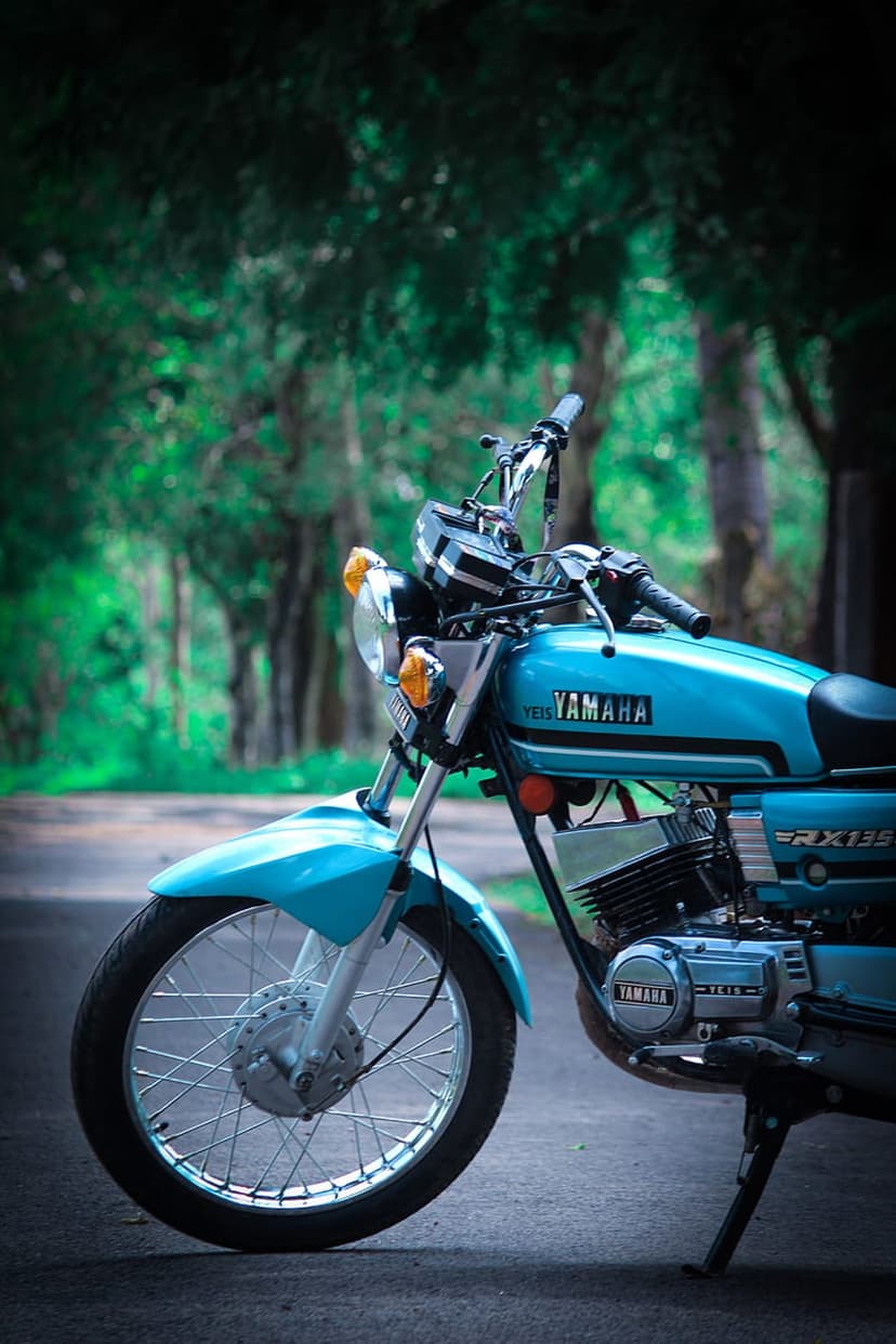 Blue Yamaha RX 135 motorcycle parked on a forest road with chrome accents and spoked wheels in soft light.
