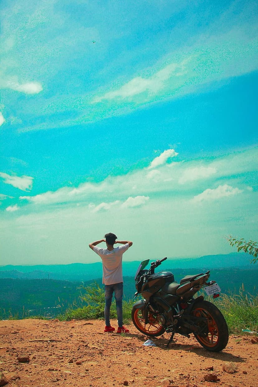 Man stands by a black motorcycle looking at a scenic mountain valley under a blue sky with scattered clouds.
