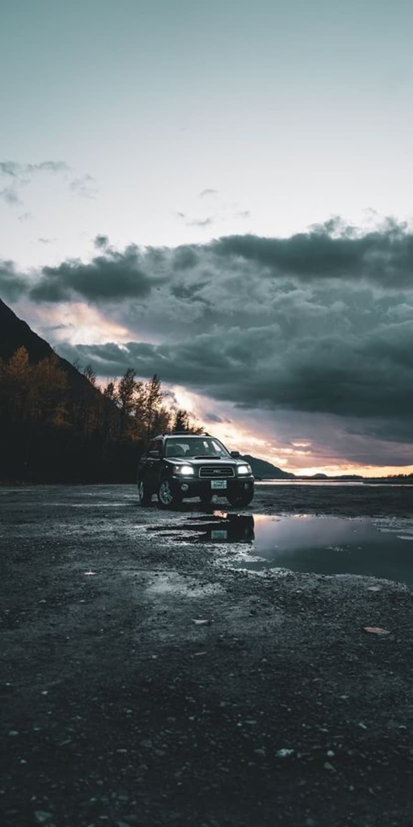 Subaru Forester SUV parked on a gravel shore by a calm mountain lake reflecting a dramatic sunset sky.