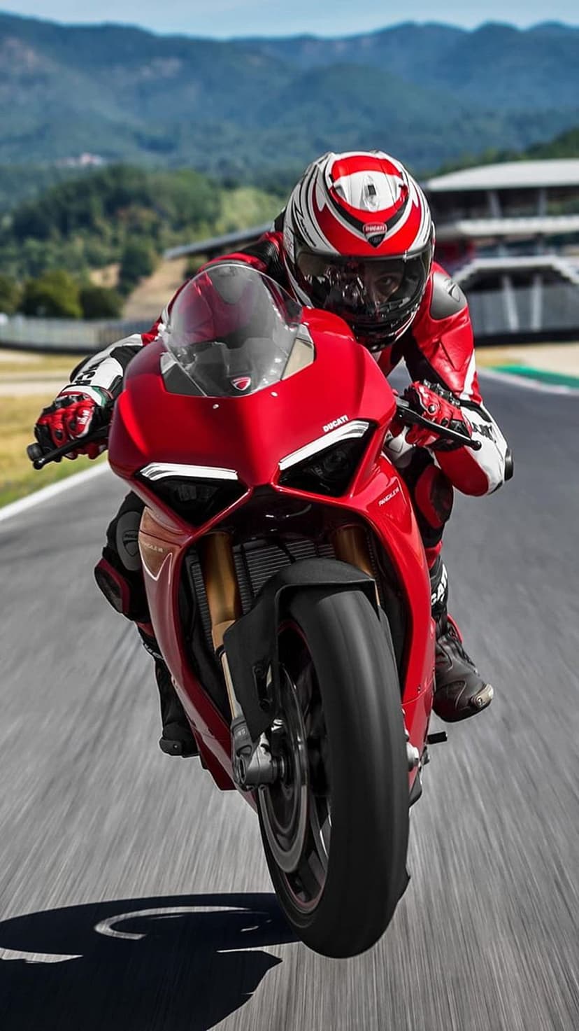 Red Ducati motorcycle racer leaning deep into a turn on a paved mountain race track under a clear blue sky.