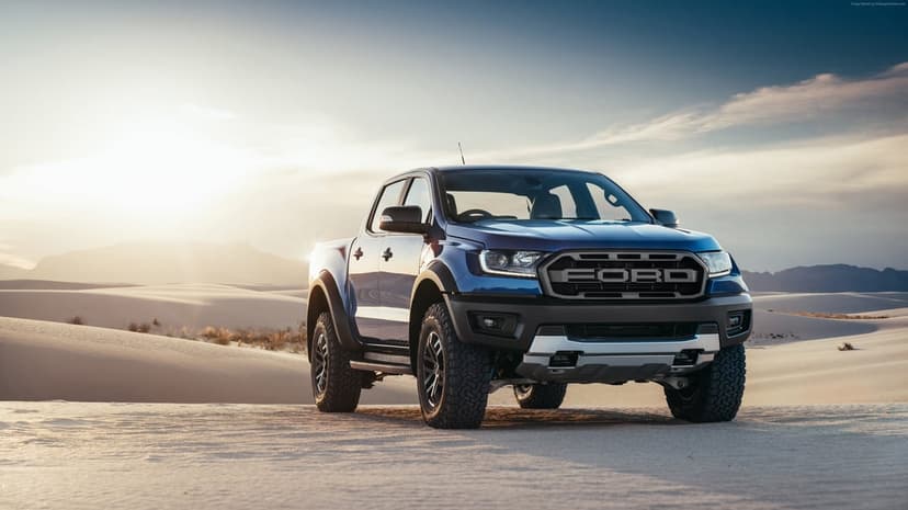 Blue Ford Ranger Raptor pickup truck parked on white sand dunes at sunset with a hazy sky background.
