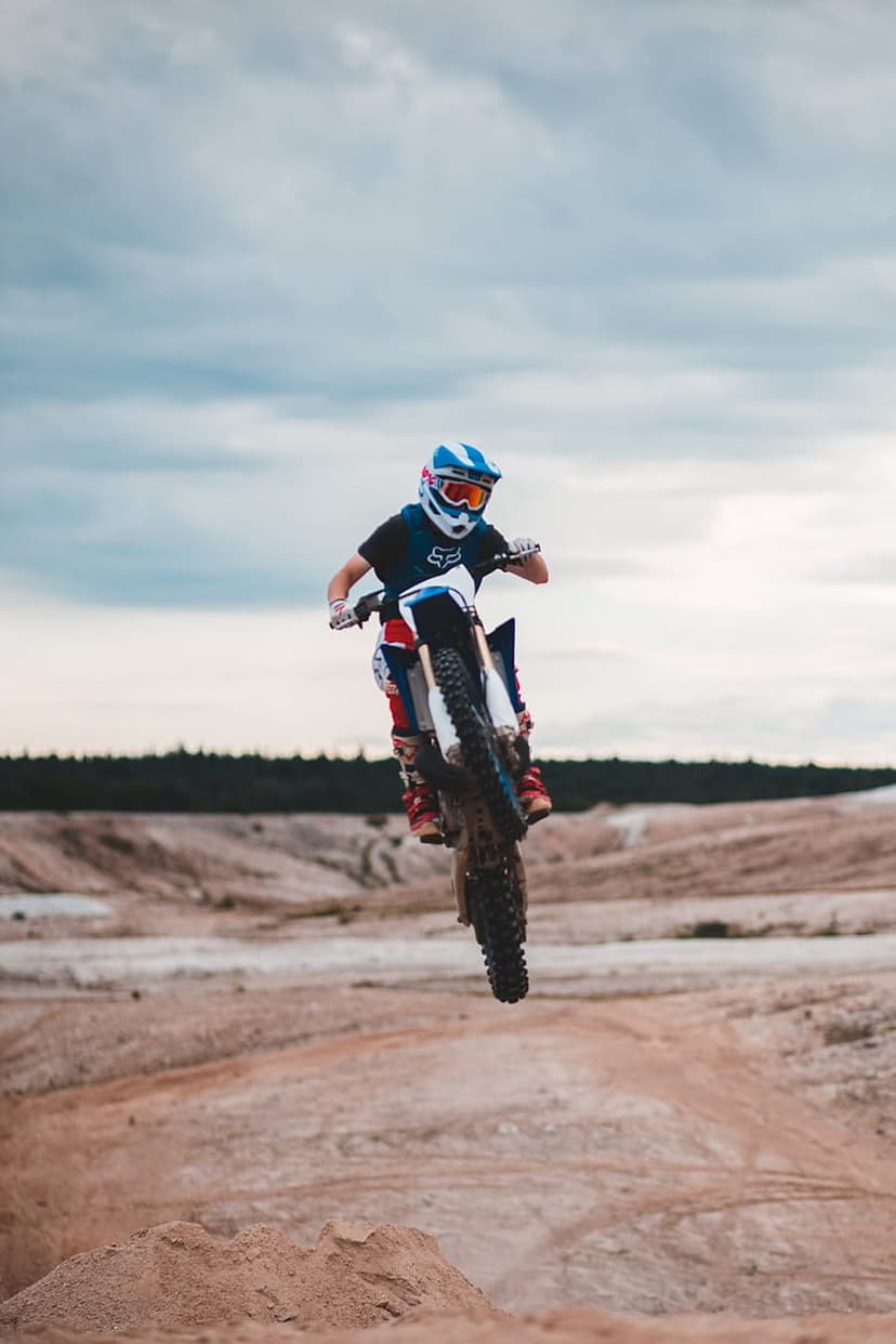 Motocross rider in blue helmet jumping a dirt bike over a sandy track under a dramatic, cloudy summer sky.