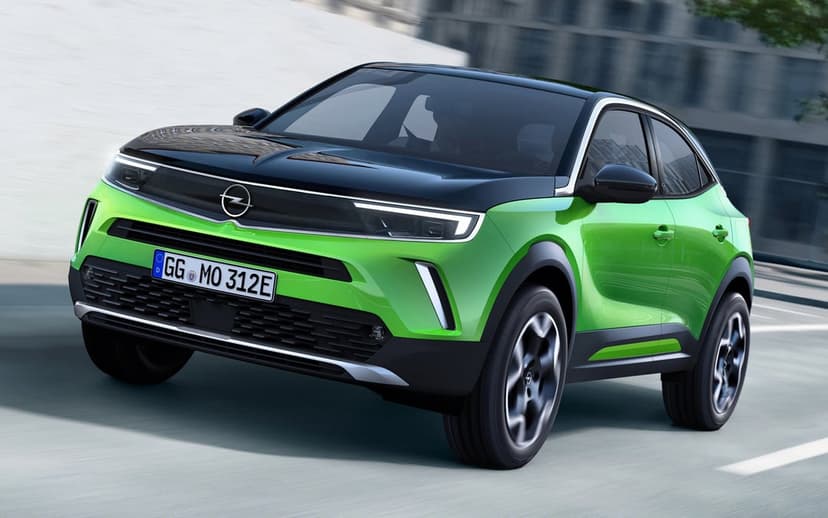 Green Opel Mokka-e electric SUV driving on a city road with a black hood and motion-blurred wheels.