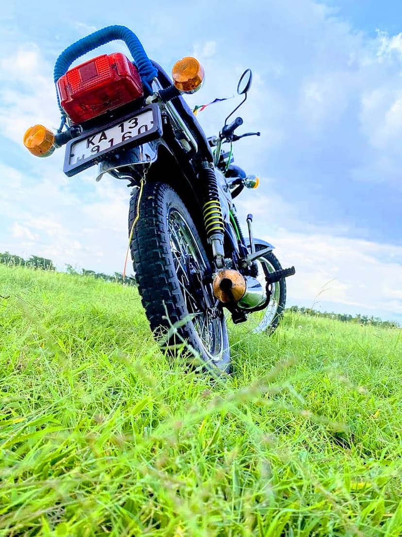 Vintage motorcycle with a red tail light and chrome accents parked in a tall green grass field under blue clouds.