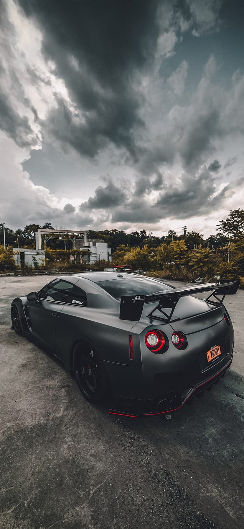 Matte black Nissan GT-R with a large spoiler parked on asphalt under a cloudy sky near an abandoned building.