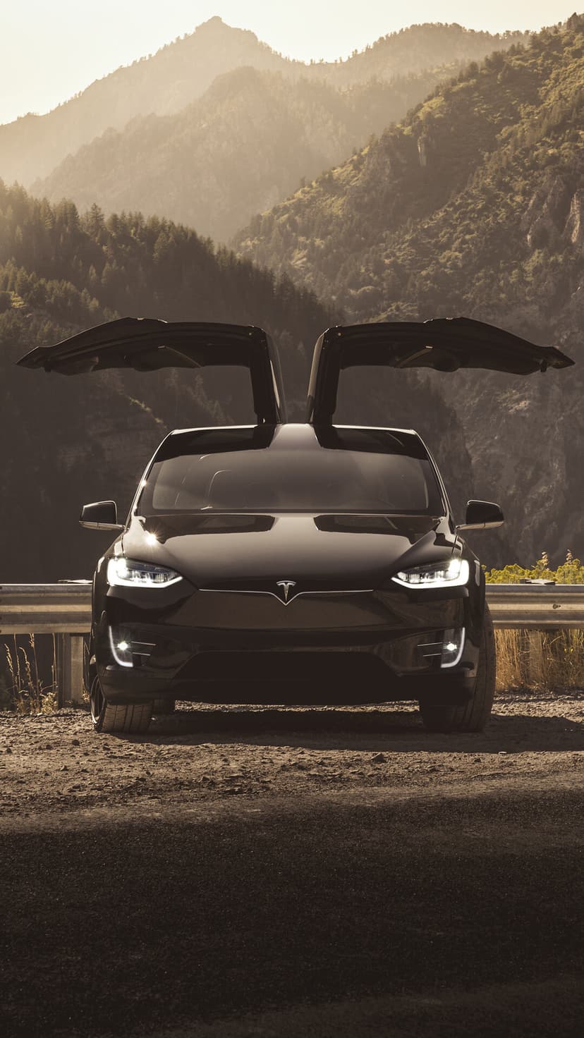 Black Tesla Model X with falcon wing doors open on a mountain road at sunset with glowing headlights.