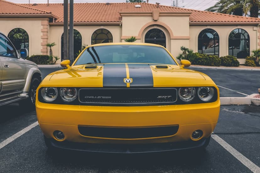 Yellow Dodge Challenger SRT with a black hood stripe and round headlights parked near palm trees and a building.