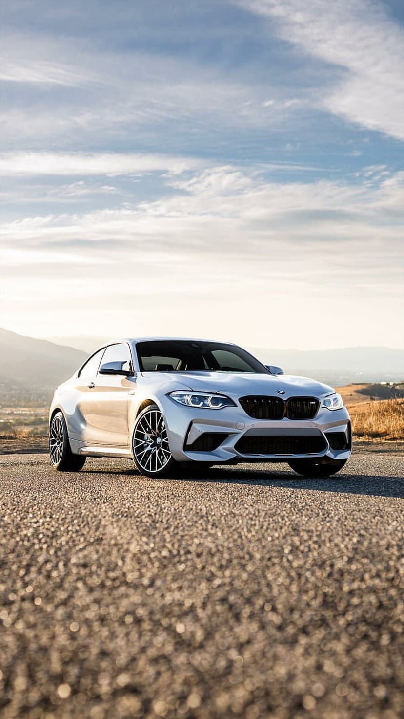 Silver BMW M2 Coupe parked on a gravel mountain road against a backdrop of distant peaks and a dramatic sky.