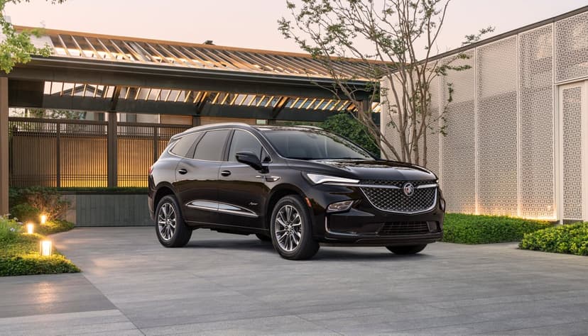 Dark brown luxury SUV parked on a paved driveway in front of a modern house with warm lighting at sunset.