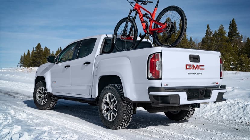 White GMC Canyon AT4 truck carrying a red mountain bike through a bright snowy landscape with a blue sky.