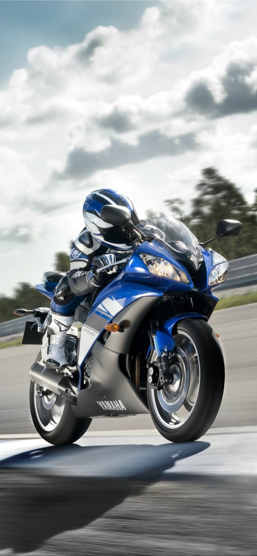 Blue Yamaha R6 motorcycle with a geared rider racing on a track under a cloudy sky with motion blur.