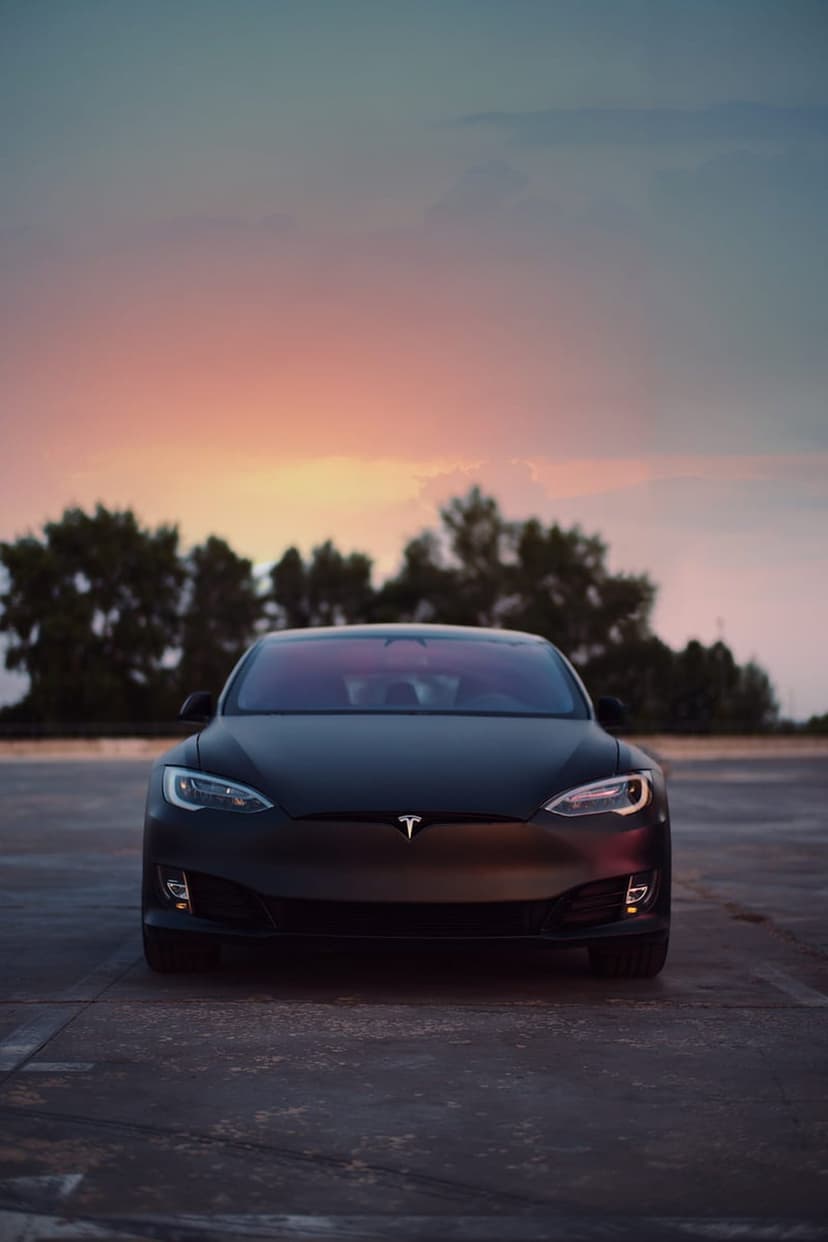 Tesla Model S in matte black finish on dark asphalt against a glowing sunset sky with distant tree silhouettes.