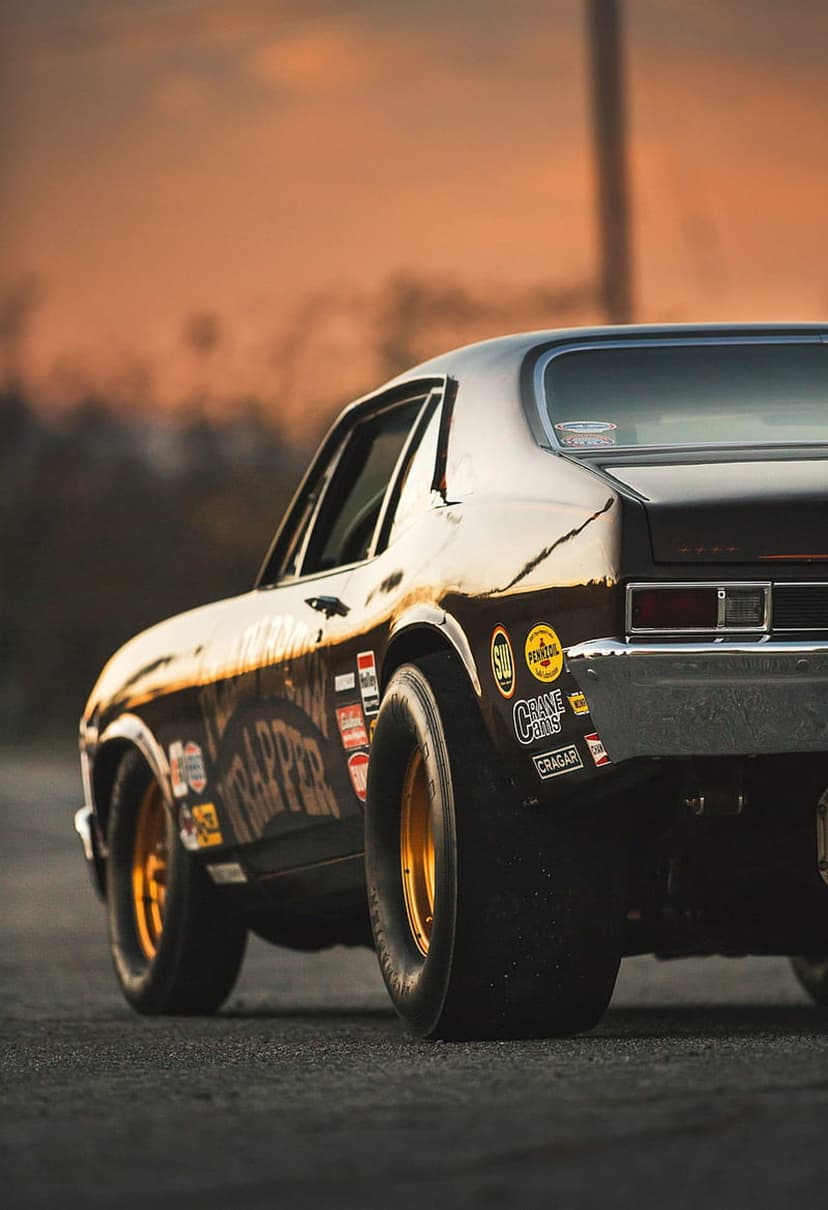 Black Trapper drag car with golden rims and sponsor decals sits under a warm sunset sky in a low-angle view.