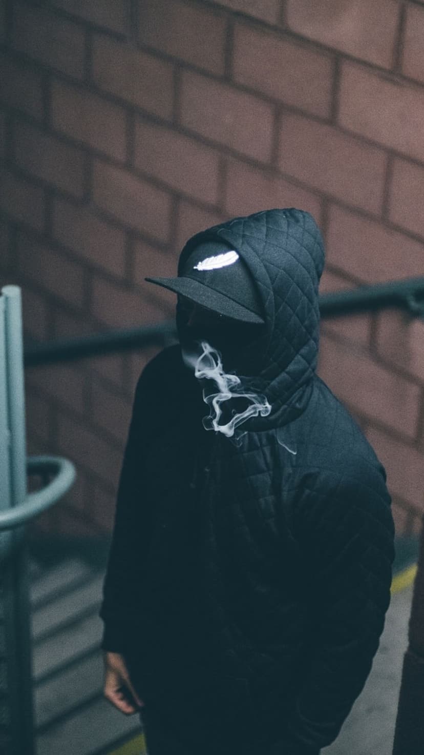 Hooded figure with glowing X eyes stands near a rainy street and a person holding a flaming newspaper outdoors.