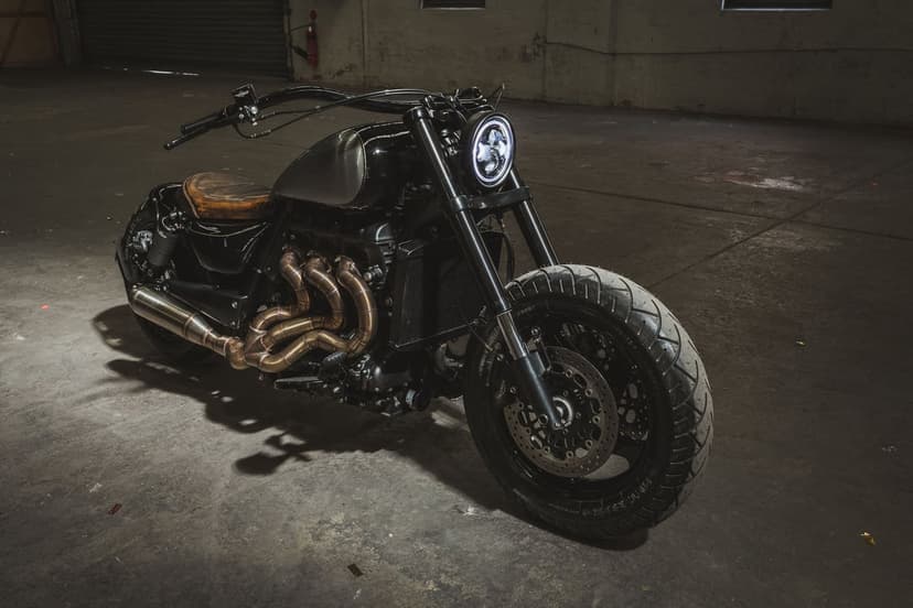 Black custom motorcycle with bronze exhaust pipes and a round LED headlight in a dark industrial warehouse.