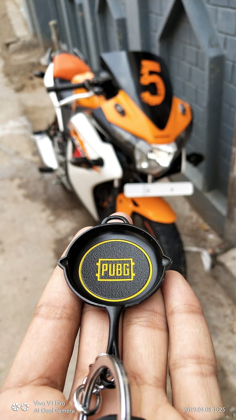 Person holding a black PUBG frying pan keychain with a blurred orange and white motorcycle in the background.