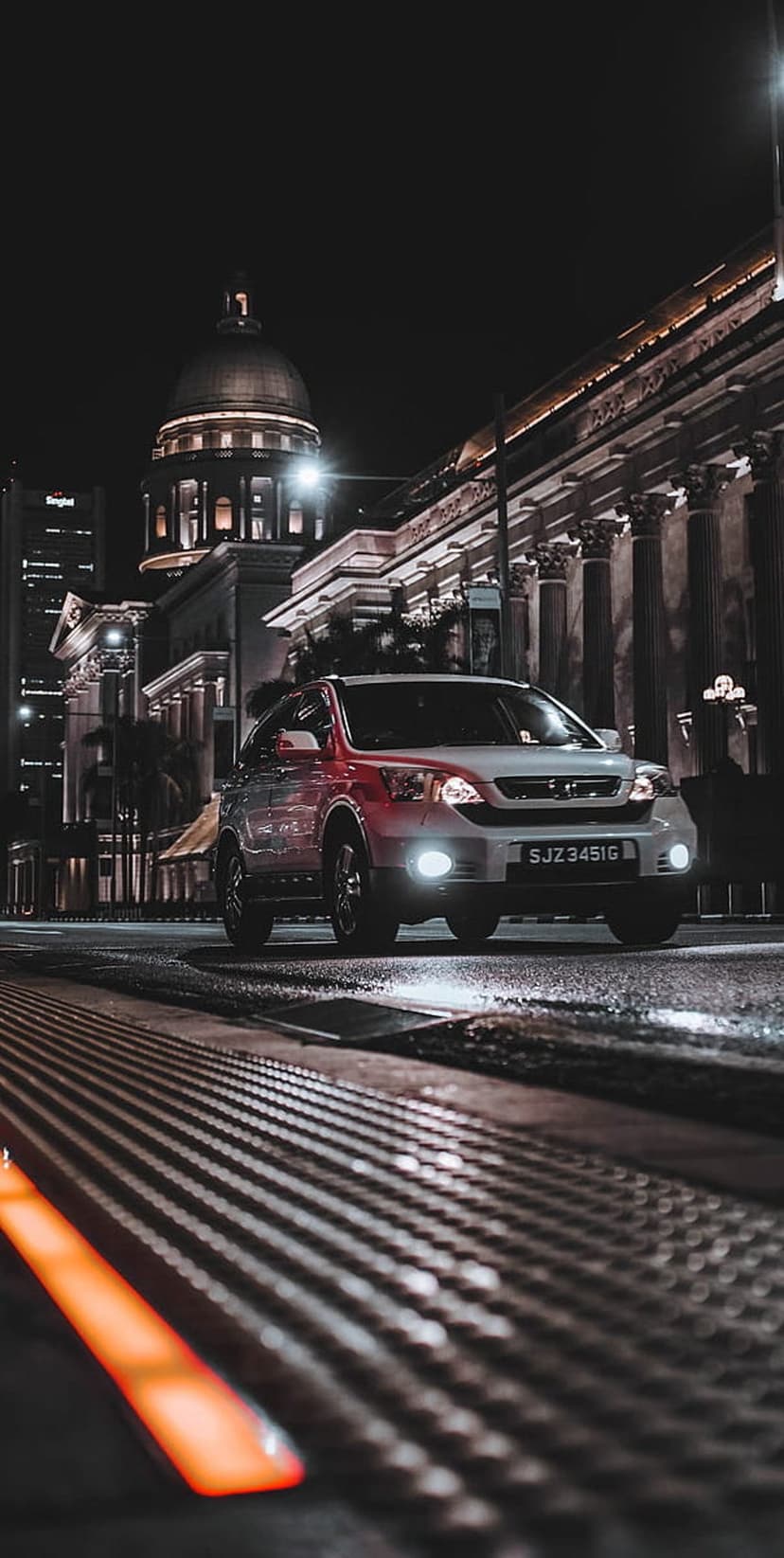 White SUV on wet asphalt in front of a lit historic dome building with orange neon light in the foreground.