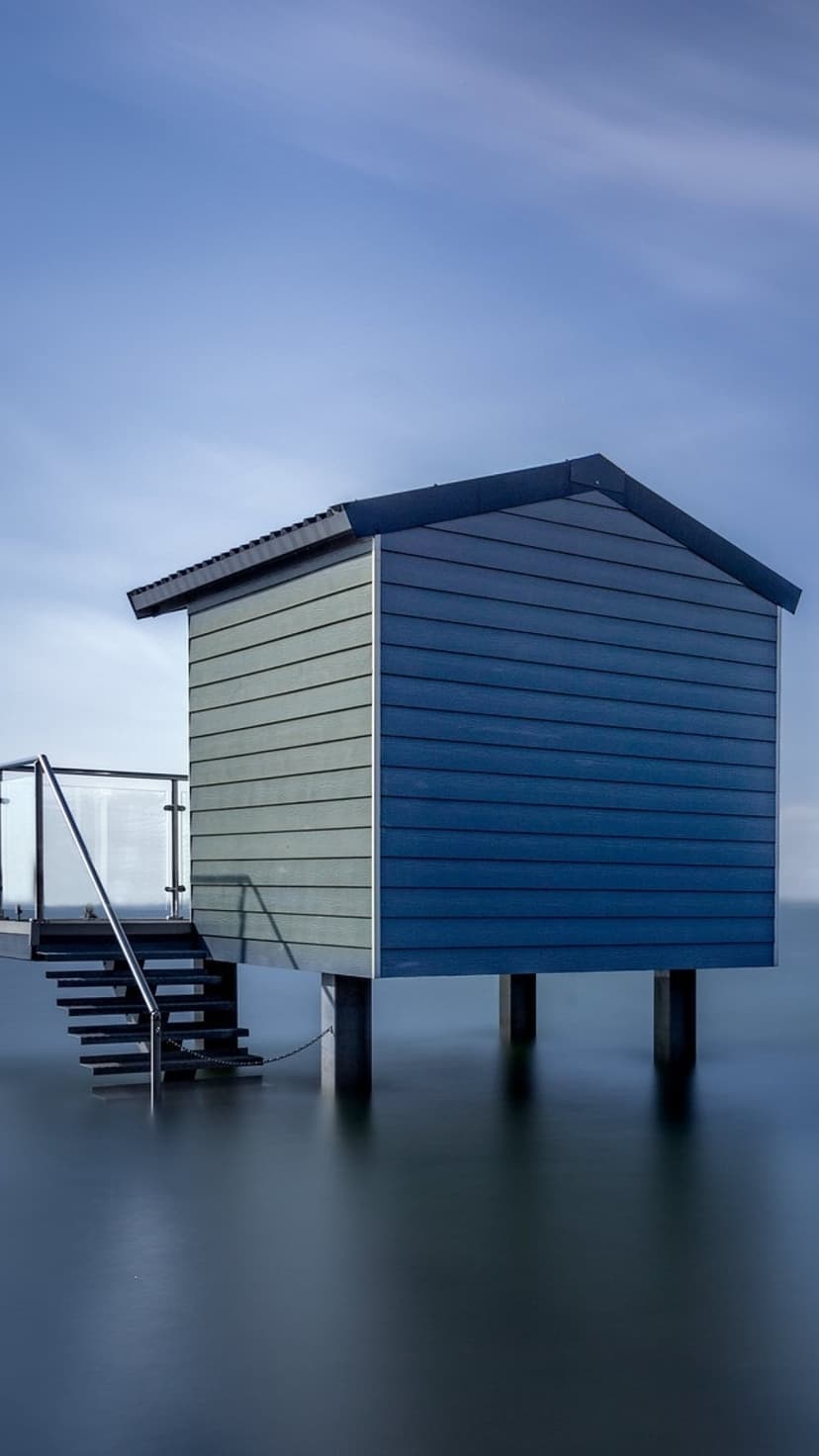 Modern two-tone beach house on stilts above still water with a glass staircase under a clear blue sky background.