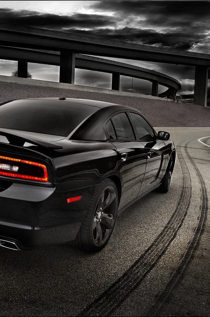 Black Dodge Charger muscle car spins on asphalt with smoke and tire tracks against a dark, dramatic cloudy sky.