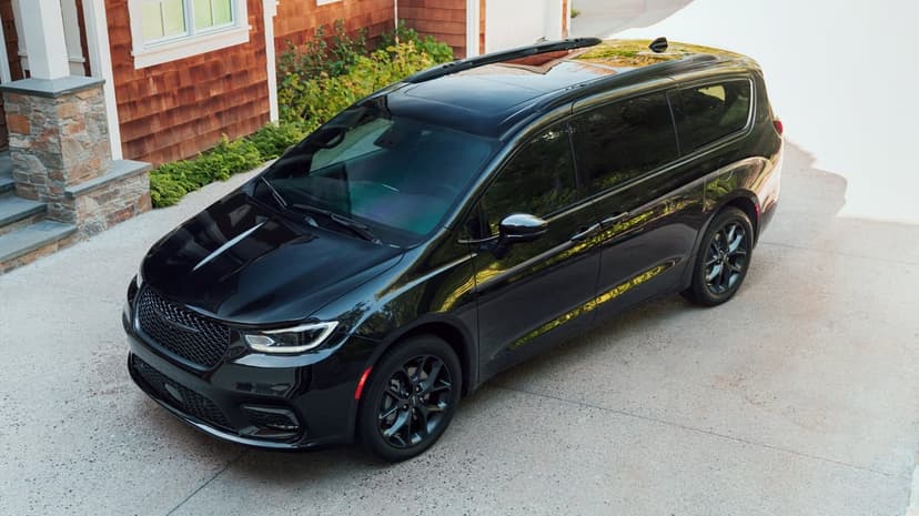 Black Chrysler Pacifica parked on a concrete driveway beside a brown shingle house under bright daylight.
