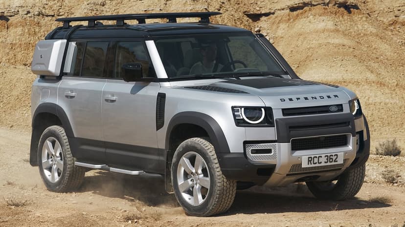 Silver Land Rover Defender driving on a sandy trail with dust kicking up around tires and a rocky backdrop.