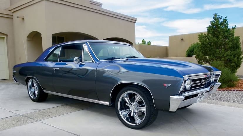 1967 Chevrolet Chevelle with blue and gray custom paint and chrome wheels parked in front of a stucco house.