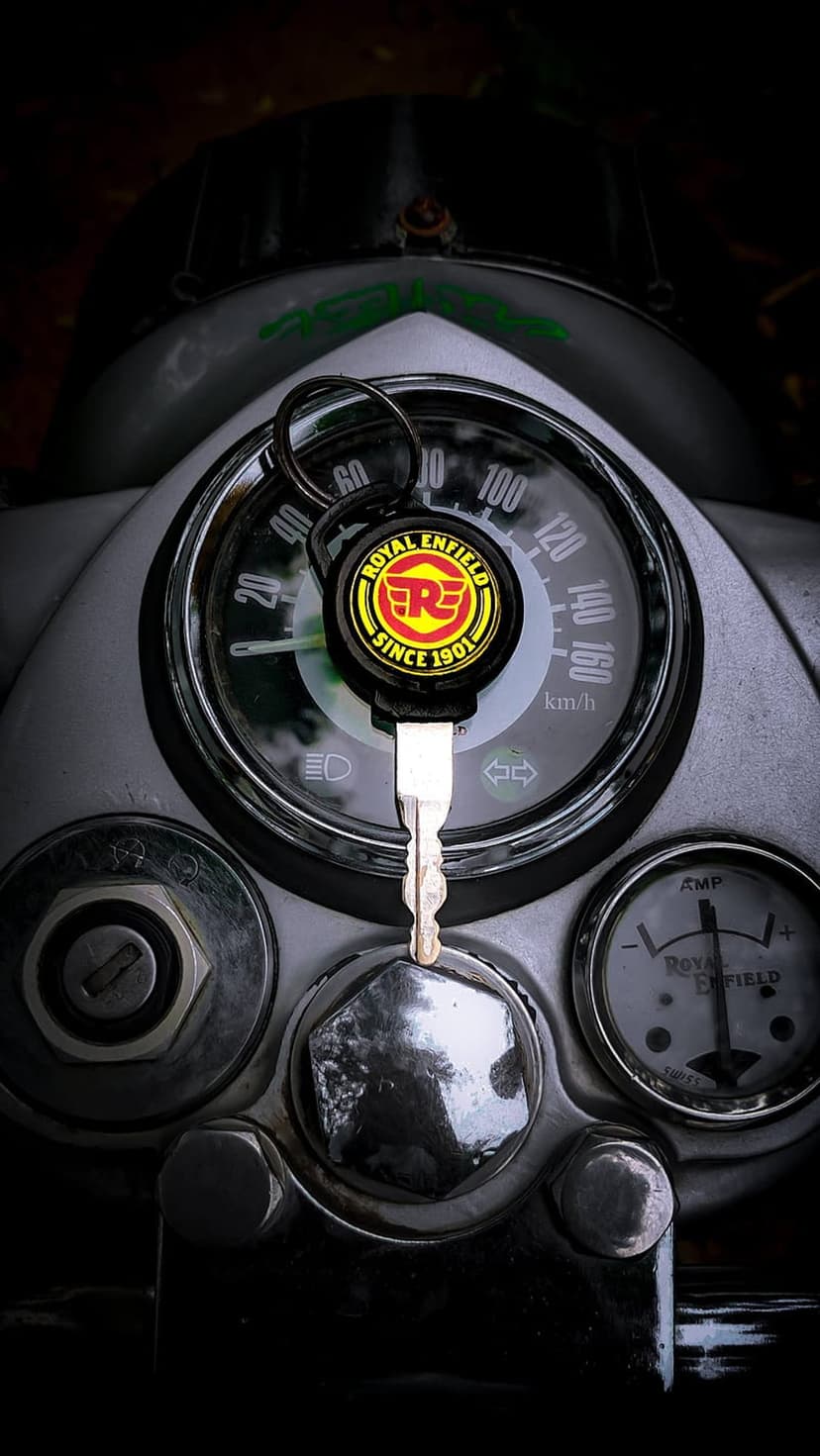 Royal Enfield ignition key inserted in a chrome dashboard with an analog speedometer under dramatic lighting.