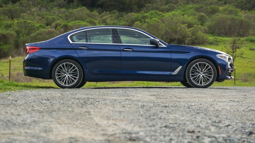Blue BMW 5 Series sedan parked on a grey gravel path with green hills and trees in the background.