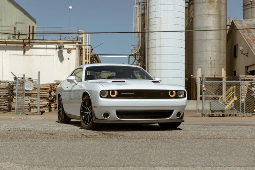 White Dodge Challenger SRT Hellcat parked in a sunlit industrial lot before massive concrete silos and buildings.