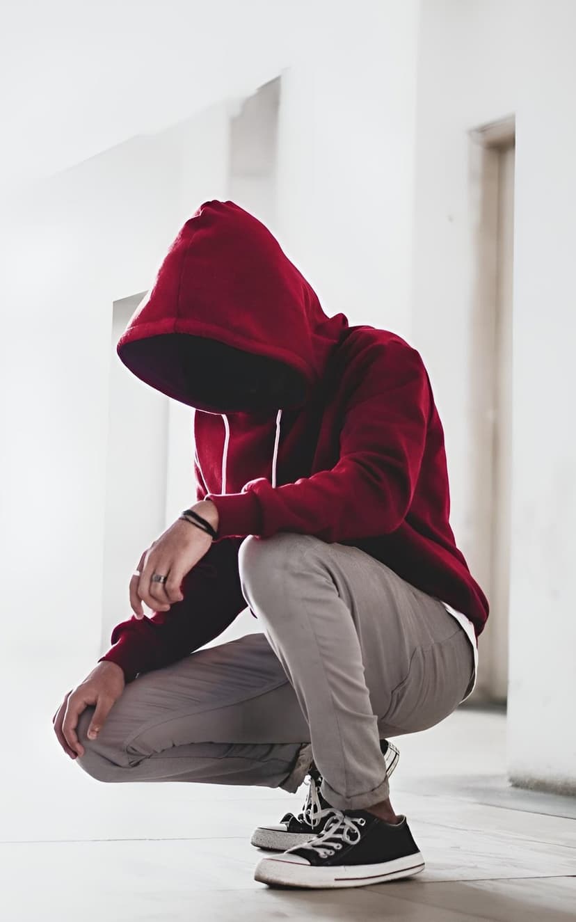 Man in a red hooded sweatshirt squats in front of a white wall wearing gray trousers and black canvas sneakers.