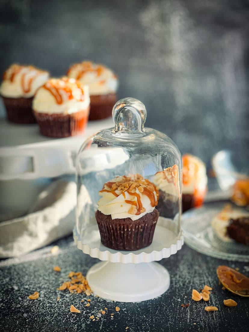Chocolate cupcake topped with white frosting and caramel sauce sits under a glass dome on a white cake stand.