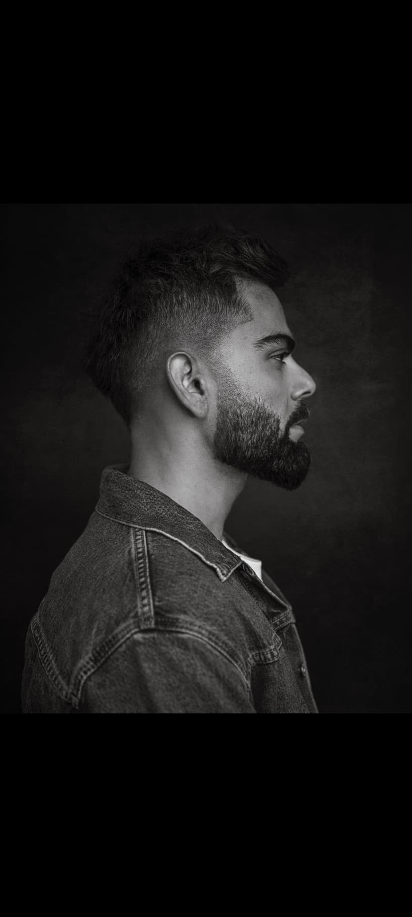 Virat Kohli shows a side profile wearing a denim jacket and groomed beard in a black and white studio portrait.