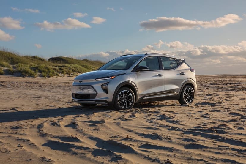Silver electric SUV parked on a sandy beach with tire tracks under a blue sky with scattered clouds.
