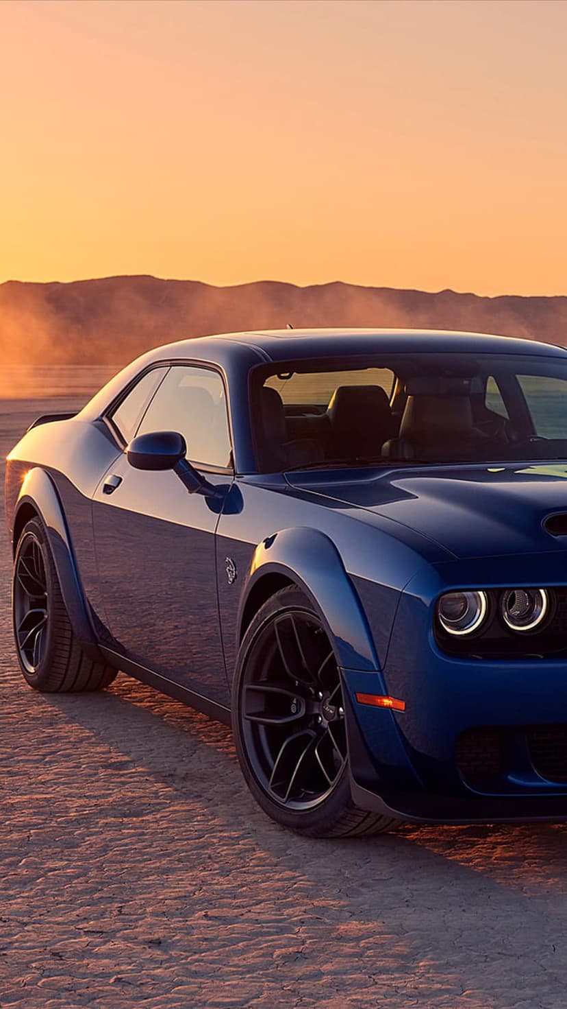 Blue Dodge Challenger with a widebody kit stands on a dusty desert floor under a golden sunset sky.
