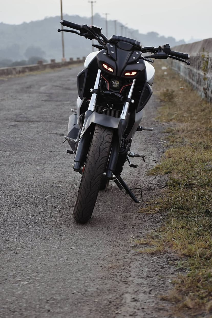 Yamaha MT-15 motorcycle with glowing LED headlights stands on a dirt road against a misty mountain backdrop.