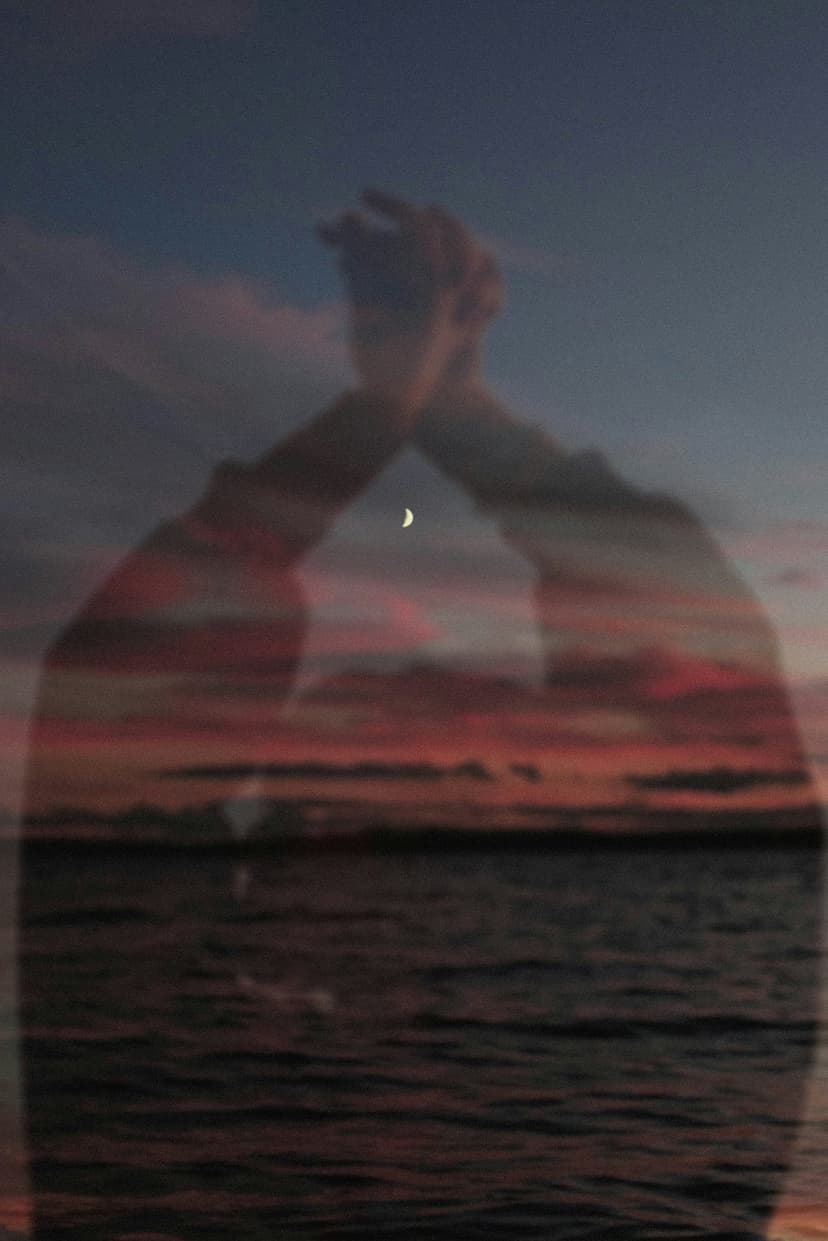 Silhouetted hands reach toward a glowing moon above dark rippling water during a vibrant orange sunset sky.