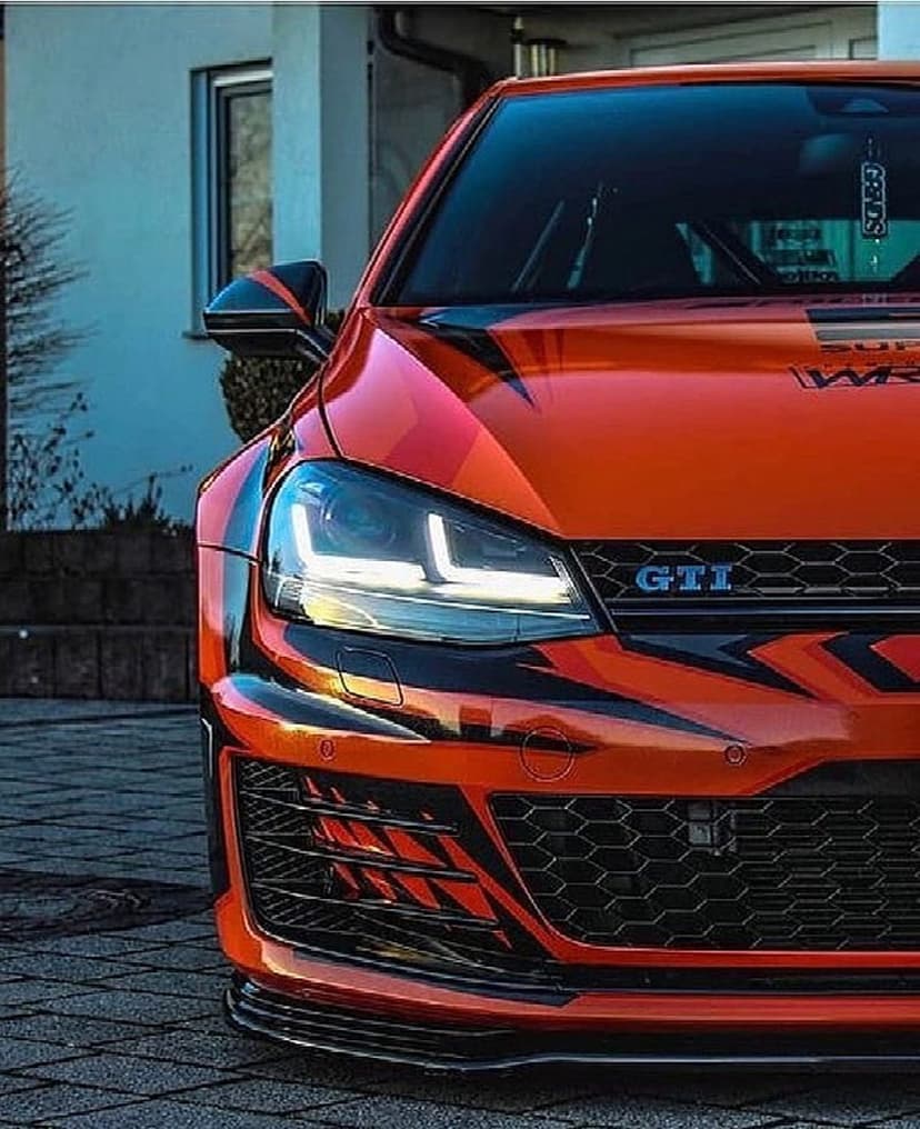 Orange Volkswagen Golf GTI with black racing stripes, glowing LED headlights, and a textured honeycomb front grille.