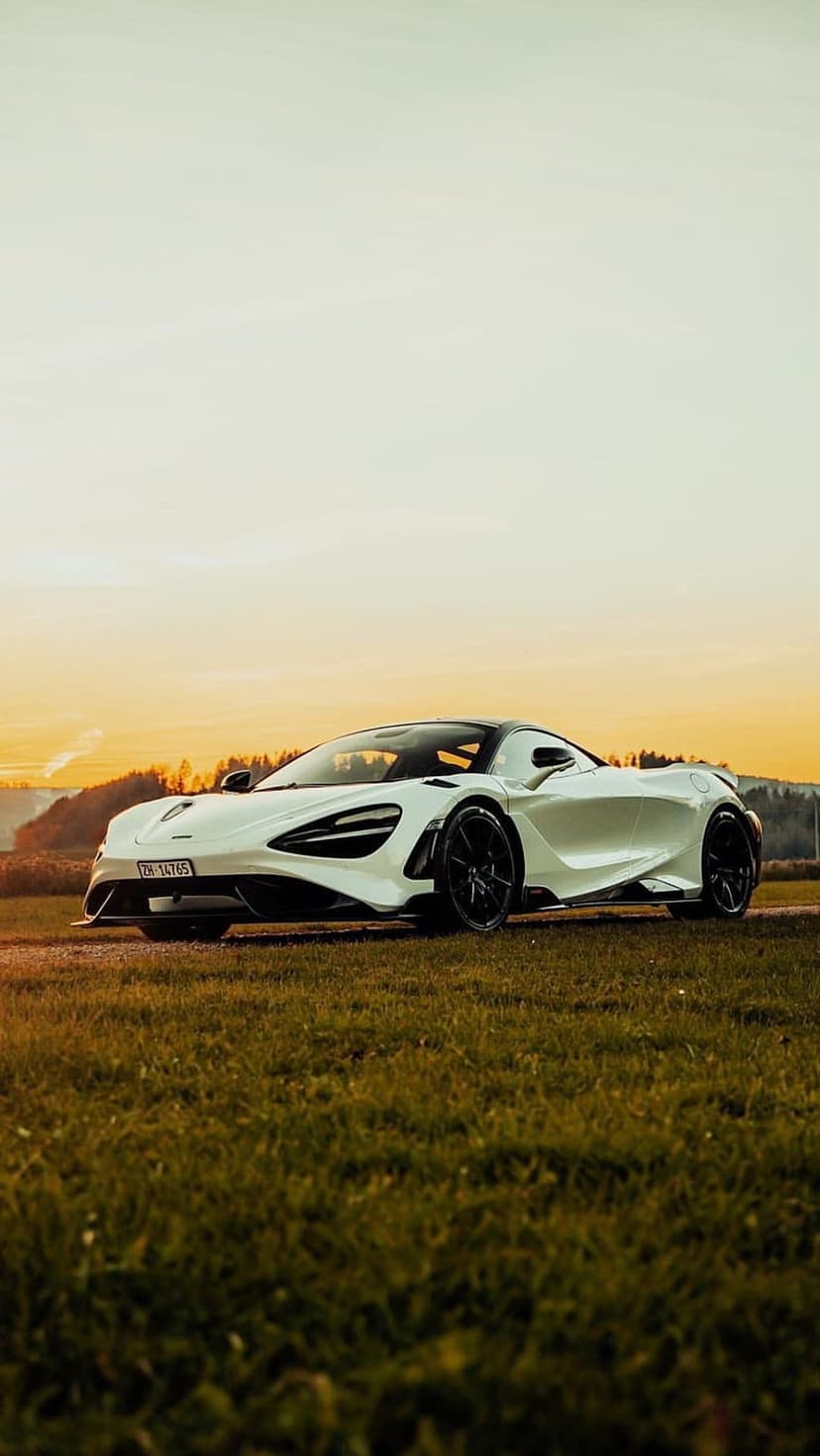 White McLaren 720S parked on a green field during a golden sunset with rolling hills in the distant background.