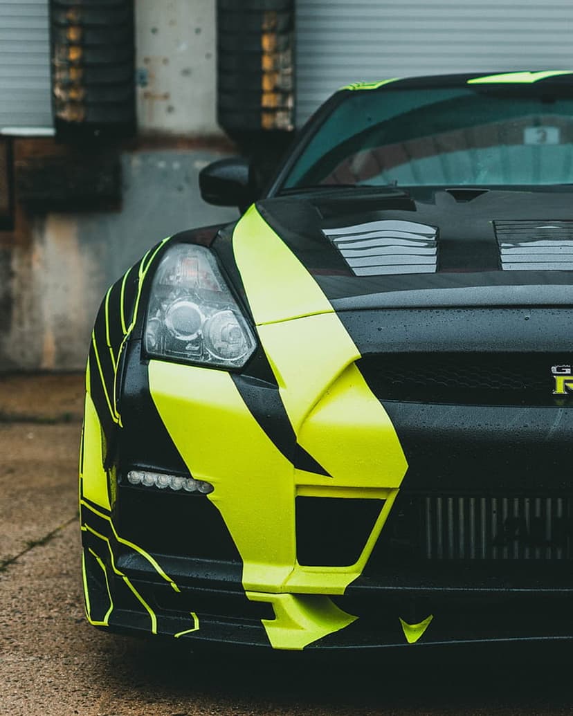 Nissan GT-R with neon yellow lightning graphics parked on concrete against a textured industrial wall backdrop.