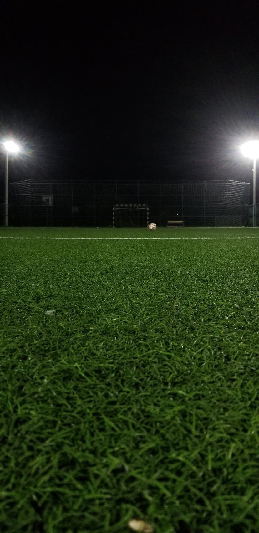 Soccer ball and goal on a floodlit artificial turf field at night under bright white stadium floodlights.
