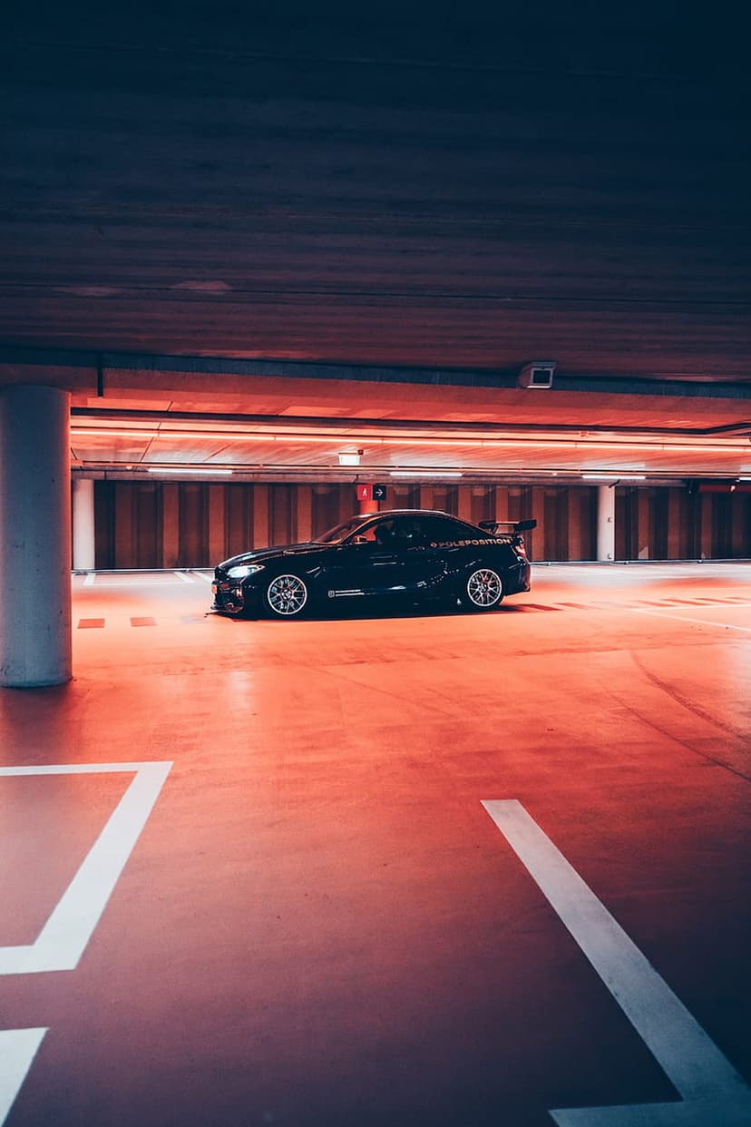 Black sports car with a rear spoiler parked in a red-lit underground garage with a reflective concrete floor.